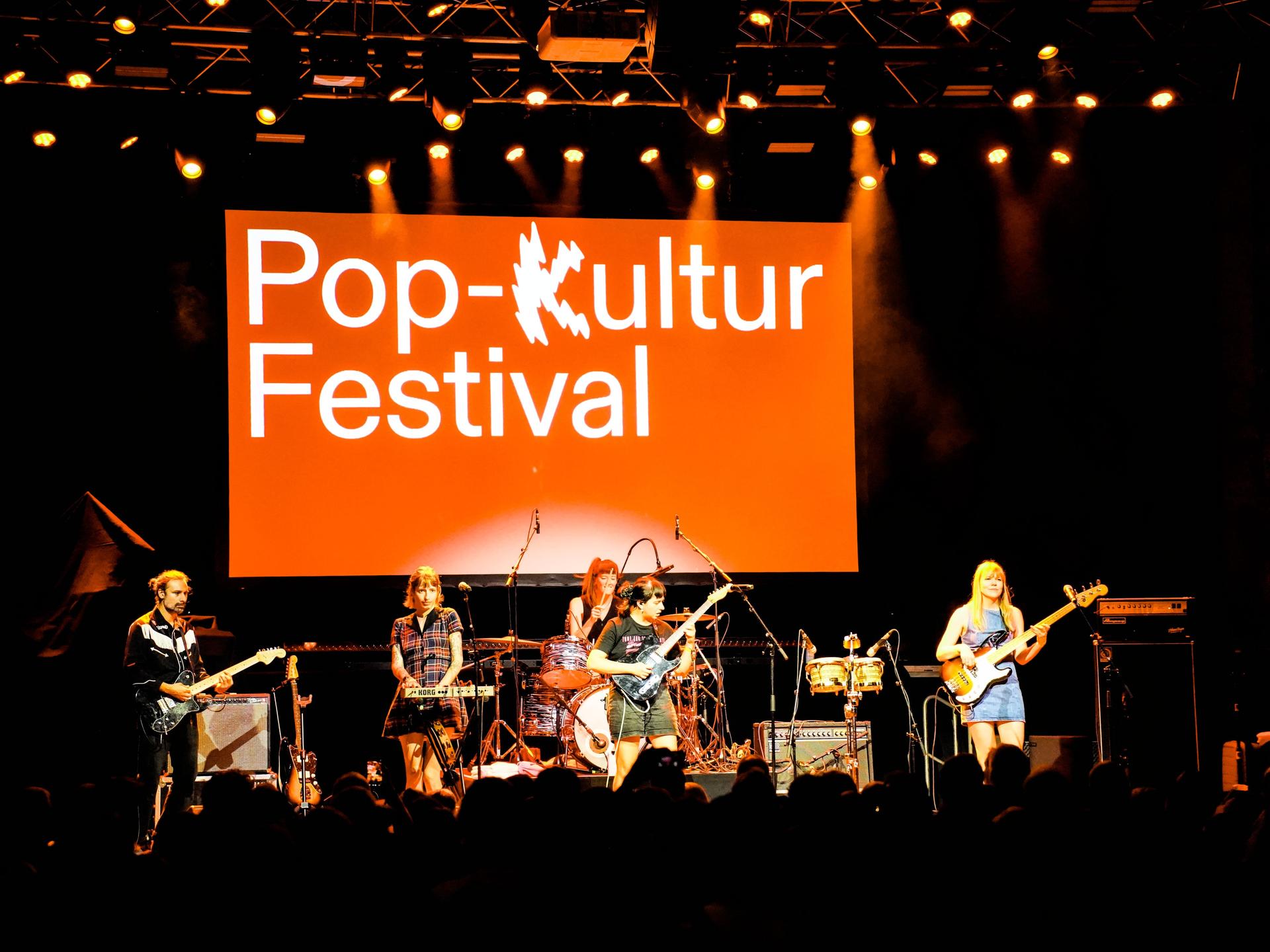 Eine Frauenband steht auf einer Bühne, hinter der auf einer orangenen Fläche "Pop-Kultur Festival" zu lesen ist.