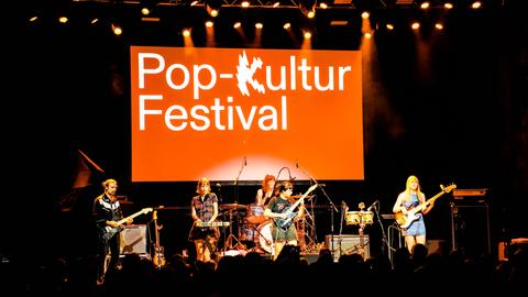 Eine Frauenband steht auf einer Bühne, hinter der auf einer orangenen Fläche "Pop-Kultur Festival" zu lesen ist.