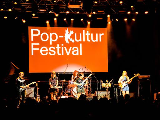 Eine Frauenband steht auf einer Bühne, hinter der auf einer orangenen Fläche "Pop-Kultur Festival" zu lesen ist.