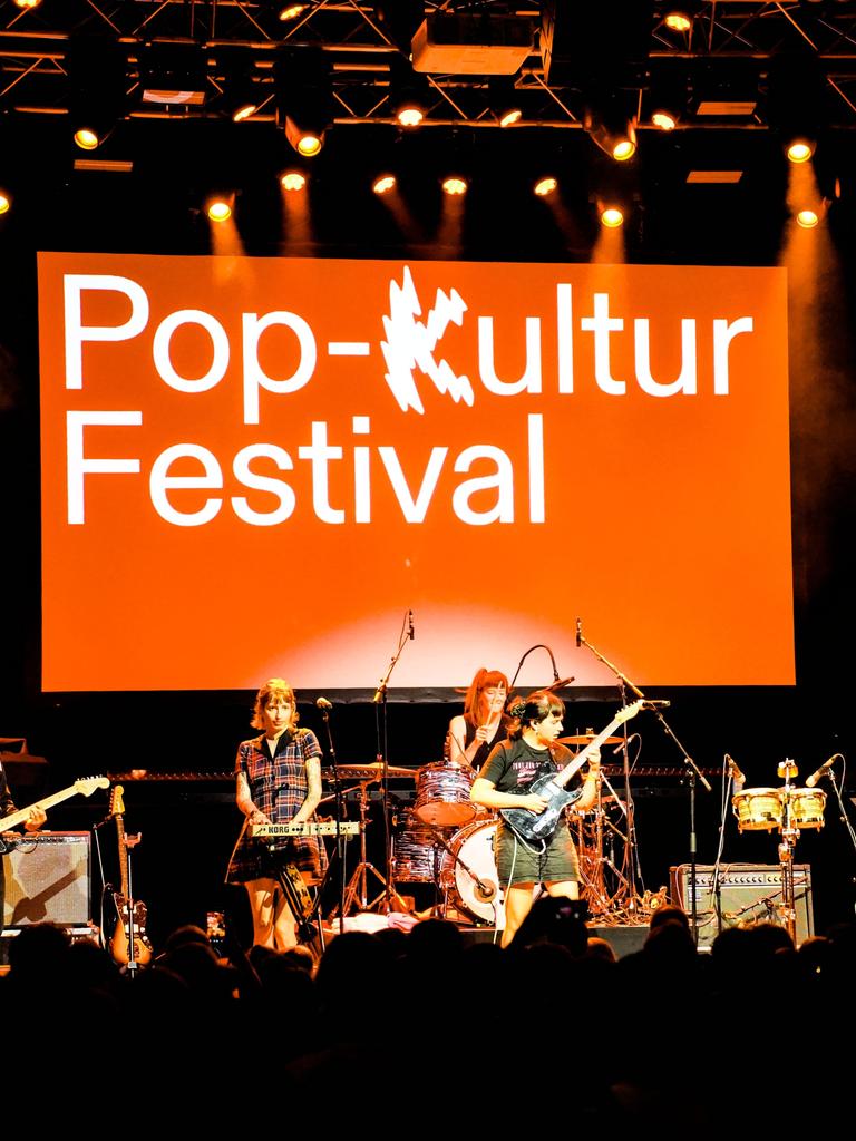 Eine Frauenband steht auf einer Bühne, hinter der auf einer orangenen Fläche "Pop-Kultur Festival" zu lesen ist.