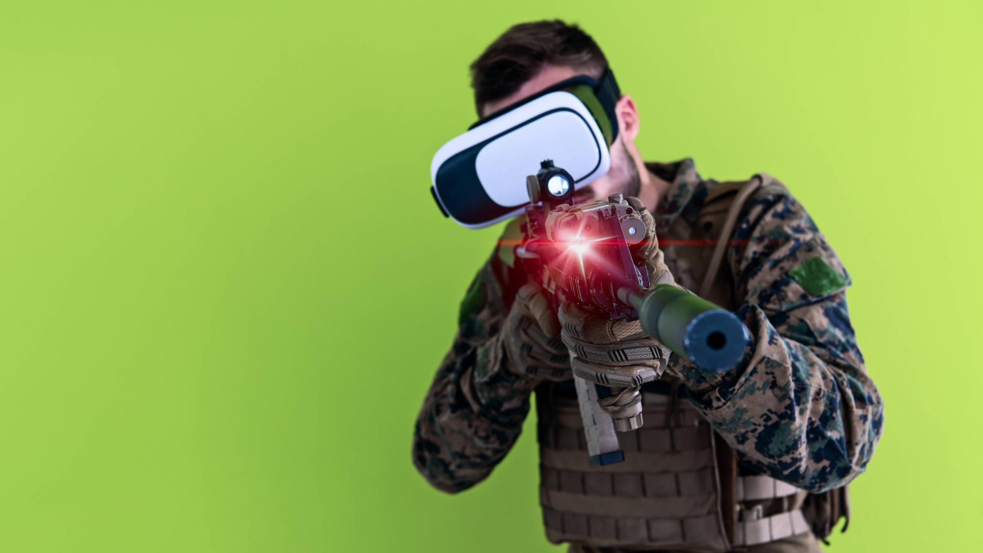 Mann in militärischer Uniform mit MG und einer Virtual Reality Brille vor grünem Hintergrund. 