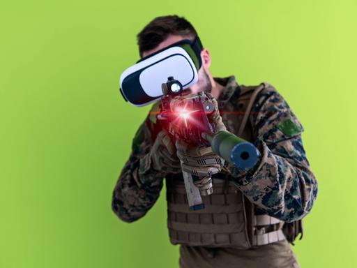 Mann in militärischer Uniform mit MG und einer Virtual Reality Brille vor grünem Hintergrund. 