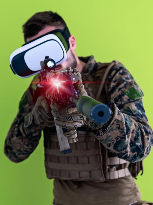 Mann in militärischer Uniform mit MG und einer Virtual Reality Brille vor grünem Hintergrund. 