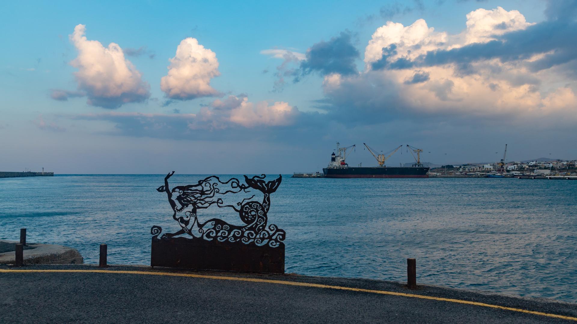 Szene an einer Uferpromenade auf Kreta. Eine Meerjungfrauinstallation ist vor dem Meer zu sehen.