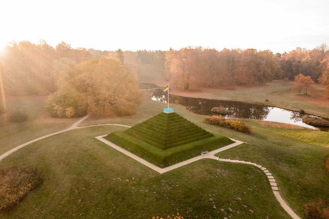 Blick von oben auf eine begrünte Steinpyramide in einem großen Park