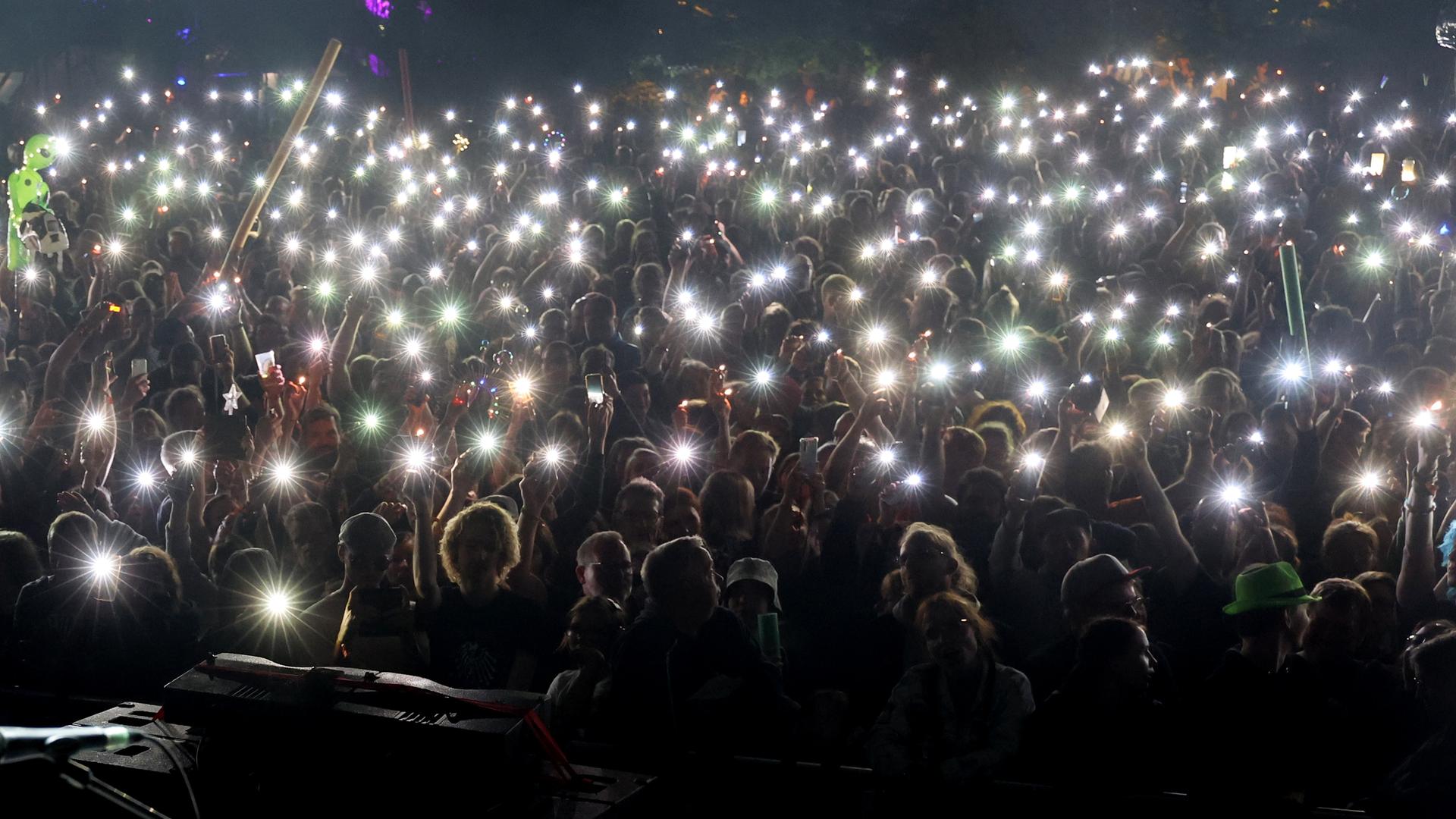 Vor einer Bühne halten viele Menschen ihre hell leuchtenden Handys ins die Dunkelheit. 