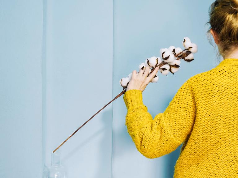 Eine Frau im gelben Pullover hält eine Baumwollpflanze in der Hand. 
