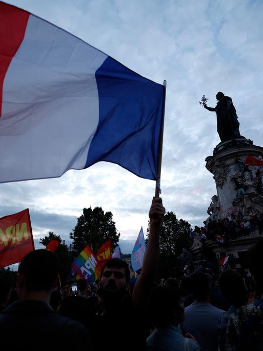 Menschen versammeln sich auf der Place de la République nach den Parlamentswahlen in Frankreich. Eine große französiche Nationalflagge weht neben der Mariannen-Statue. 