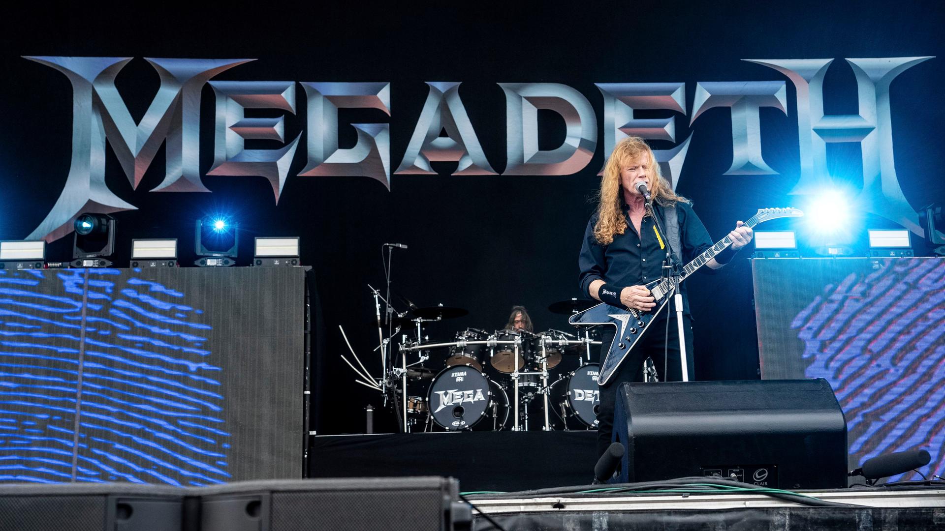 Der Sänger und Gitarrist von Megadeth, Dave Mustaine, performt auf der Bühne bei einem Festival. Hinter ihm in großen Buchstaben der Name der Band.