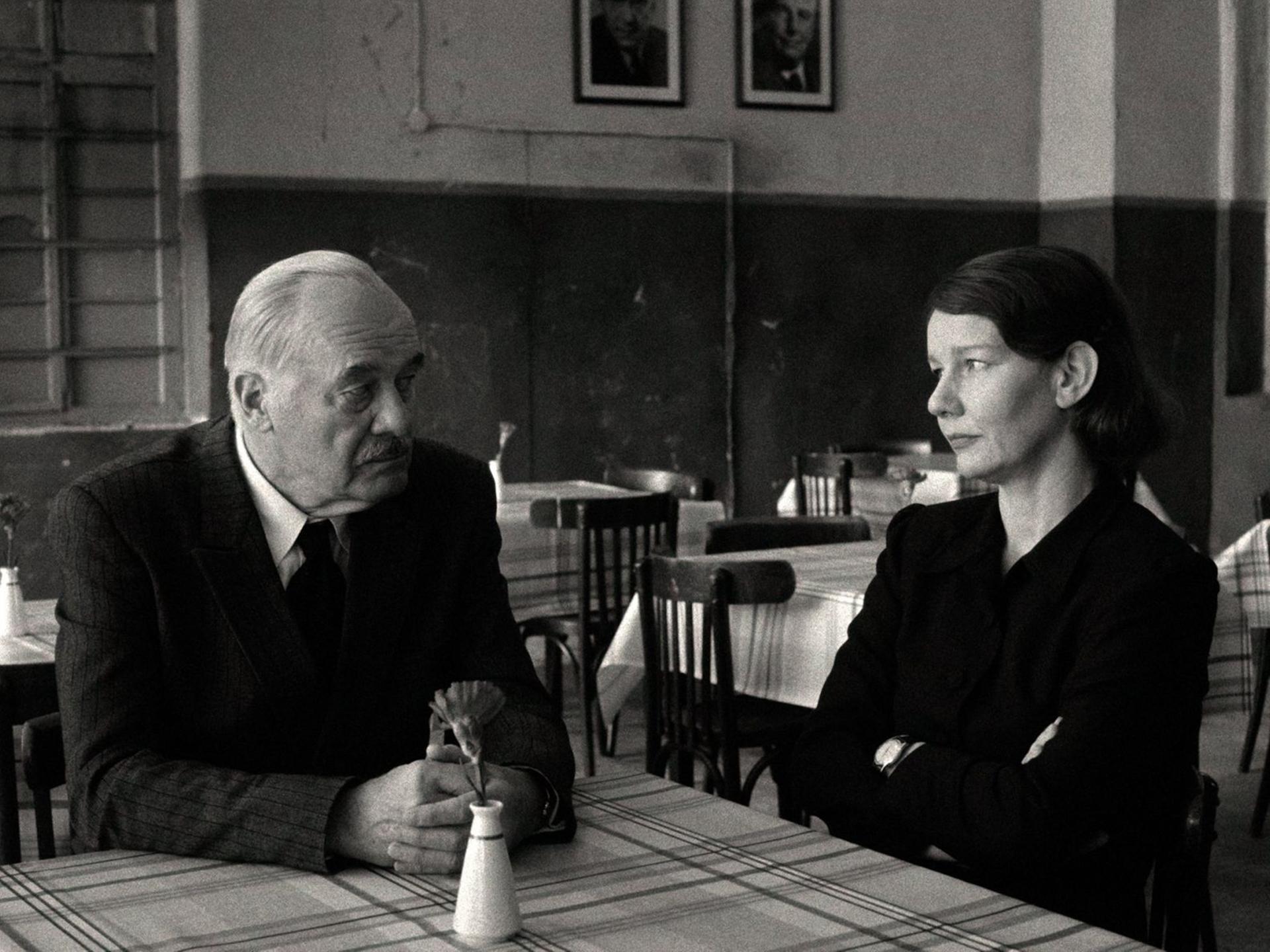 Szene aus einem Schwarz-Weiß-Film: Ein Mann und eine Frau sitzen an einem Tisch und schauen sich an.