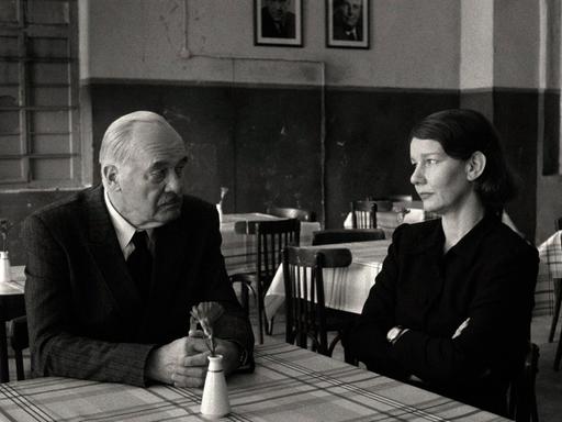 Szene aus einem Schwarz-Weiß-Film: Ein Mann und eine Frau sitzen an einem Tisch und schauen sich an.