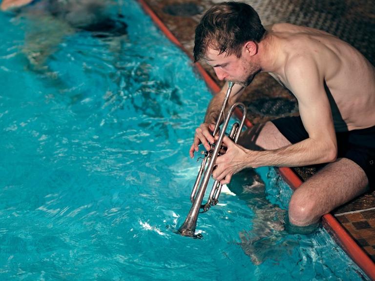 Ein Trompeter in Badehose sitzt am Schwimmbeckenrand und bespielt das Wasser