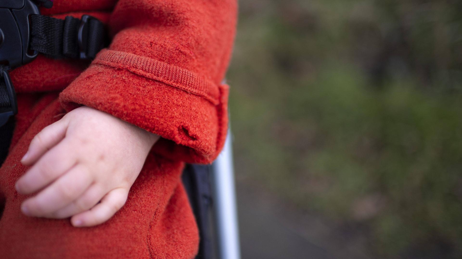 Eine Kinderhand schaut aus einem roten Mantelärmel. Der Ärmel hat ein großes Loch.