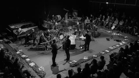 Blick auf eine Bühne mit Musikern, Dirigenten und zwei stehenden, agierenden Männern. Die Bühne ist dabei mit Efeu-Ranken umlegt. Darauf liegen diverse politische Zeitschriften.