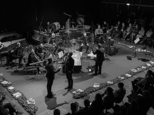 Blick auf eine Bühne mit Musikern, Dirigenten und zwei stehenden, agierenden Männern. Die Bühne ist dabei mit Efeu-Ranken umlegt. Darauf liegen diverse politische Zeitschriften. Blick auf eine Bühne mit Musikern, Dirigenten und zwei stehenden, agierenden Männern. Die Bühne ist dabei mit Efeu-Ranken umlegt. Darauf liegen diverse politische Zeitschriften.