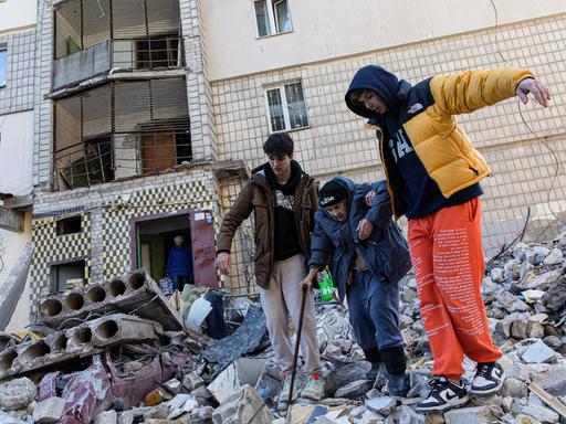 Zwei Männer helfen einer alten Frau, ein zerstörtes Gebäude zu verlassen.