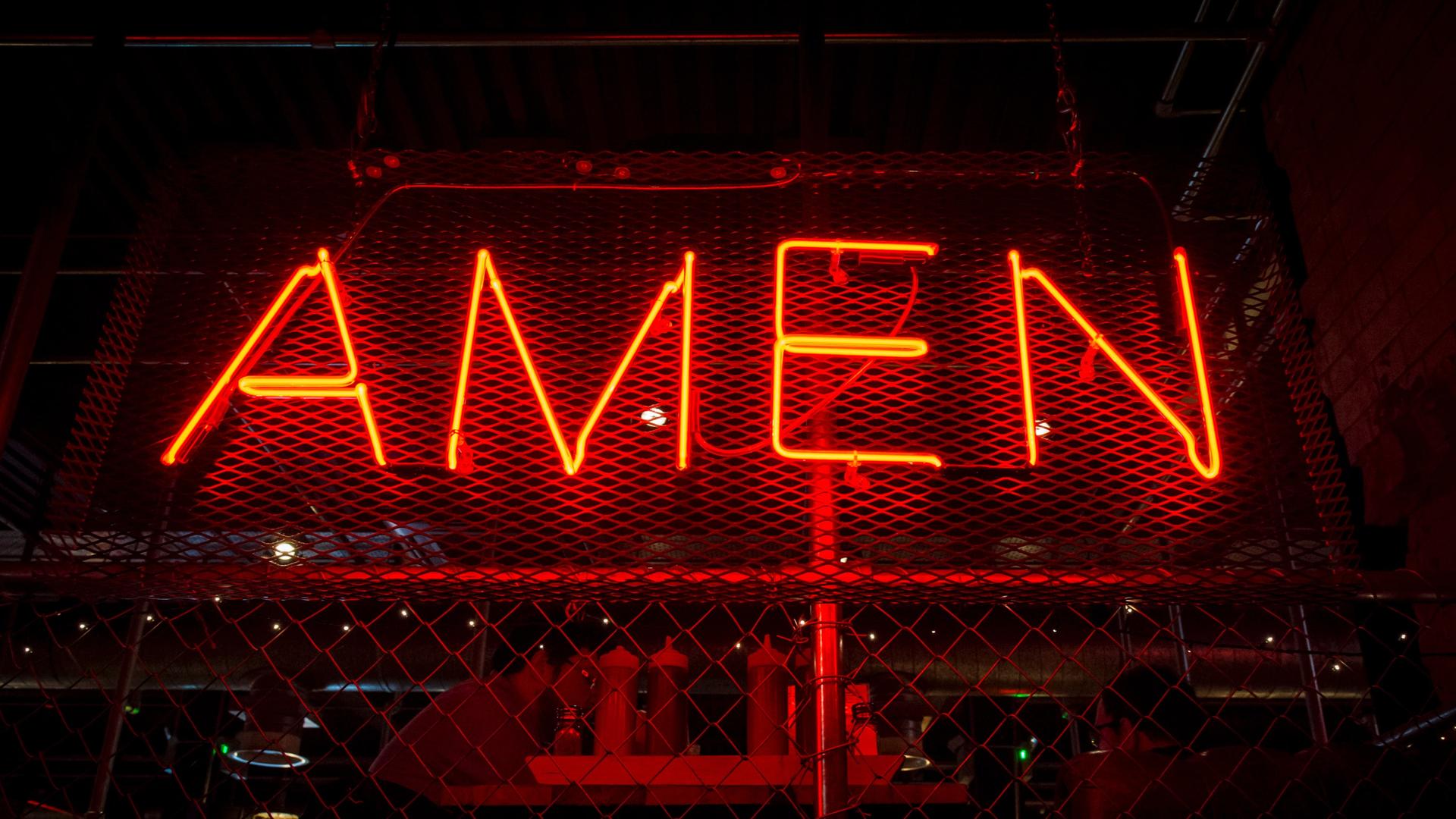In großen roten Neonbuchstaben hängt das Wort "Amen" an einem Drahtzaun.