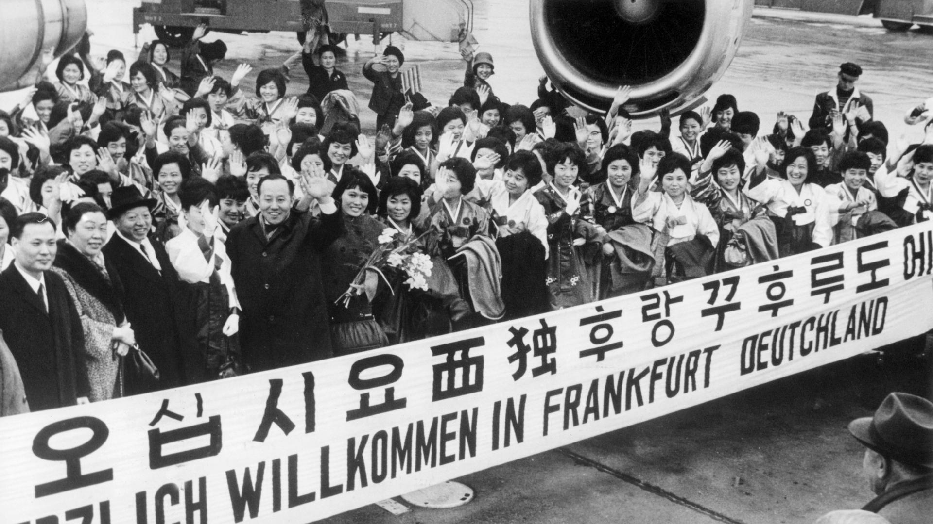 "Herzlich Willkommen in Frankfurt Deutschland" steht auf einem Spruchband, mit dem eine große Gruppe Krankenschwestern aus Südkorea im Jahr 1966 auf dem Rhein-Main-Flughafen begrüßt wird. 