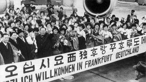 "Herzlich Willkommen in Frankfurt Deutschland" steht auf einem Spruchband, mit dem eine große Gruppe Krankenschwestern aus Südkorea im Jahr 1966 auf dem Rhein-Main-Flughafen begrüßt wird. 