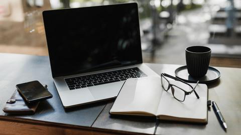 Ein Arbeitsplatz mit Laptop, Handy, Notizbuch, Stift, Brille und einer Tasse Kaffee