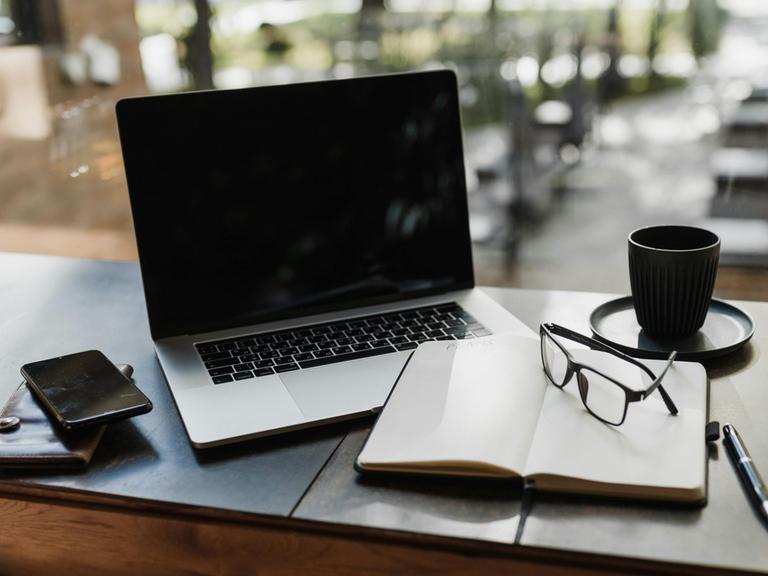 Blick auf einen Arbeitsplatz mit Laptop, Handy, Notizbuch, Stift, Brille und einer Tasse Kaffee.