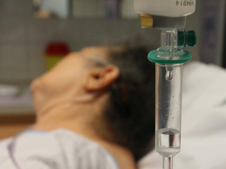 Im Vordergrund sieht man ein Edikament durch einen Tropf laufen. Im Hintergrund liegt ein Mensch im Bett mit abgewandtem Gesicht.