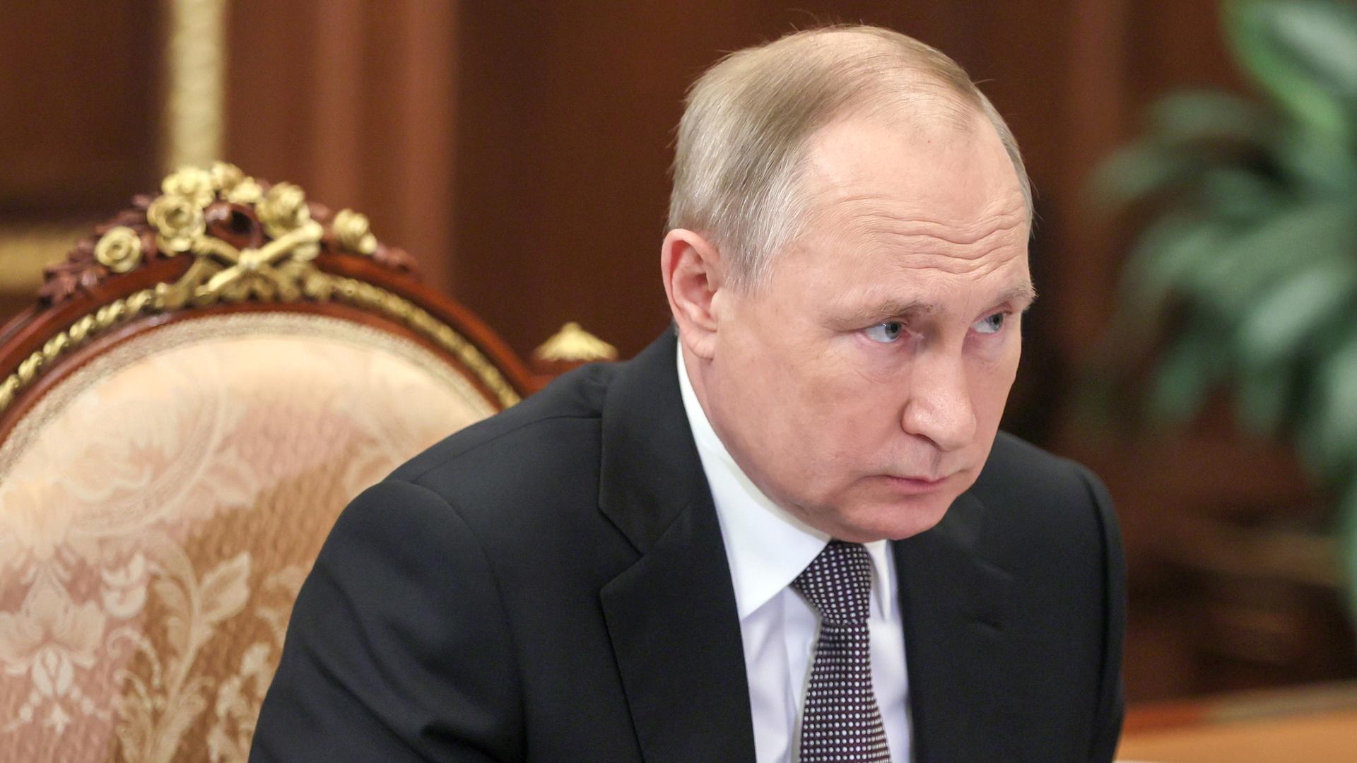 Wladimir Putin, im schwarzen Sakko mit Krawatte, sitzt auf einem Stuhl mit goldenen Ornamenten an einem Schreibtisch und neigt sich einem Gesprächspartner zu, der nicht im Bild ist.