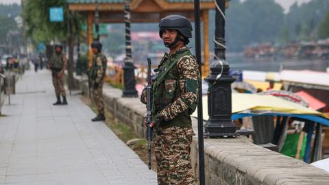 Ein Mann in militärischer Kleidung mit einem Maschinengewehr in der Hand steht an einer Promenade am Wasser. Im Hintergrund sind weitere bewaffnete Männer zu sehen.