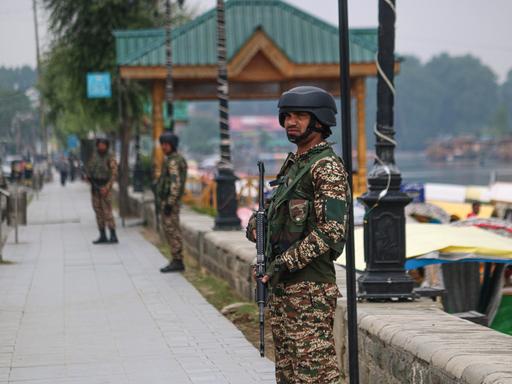 Ein Mann in militärischer Kleidung mit einem Maschinengewehr in der Hand steht an einer Promenade am Wasser. Im Hintergrund sind weitere bewaffnete Männer zu sehen.