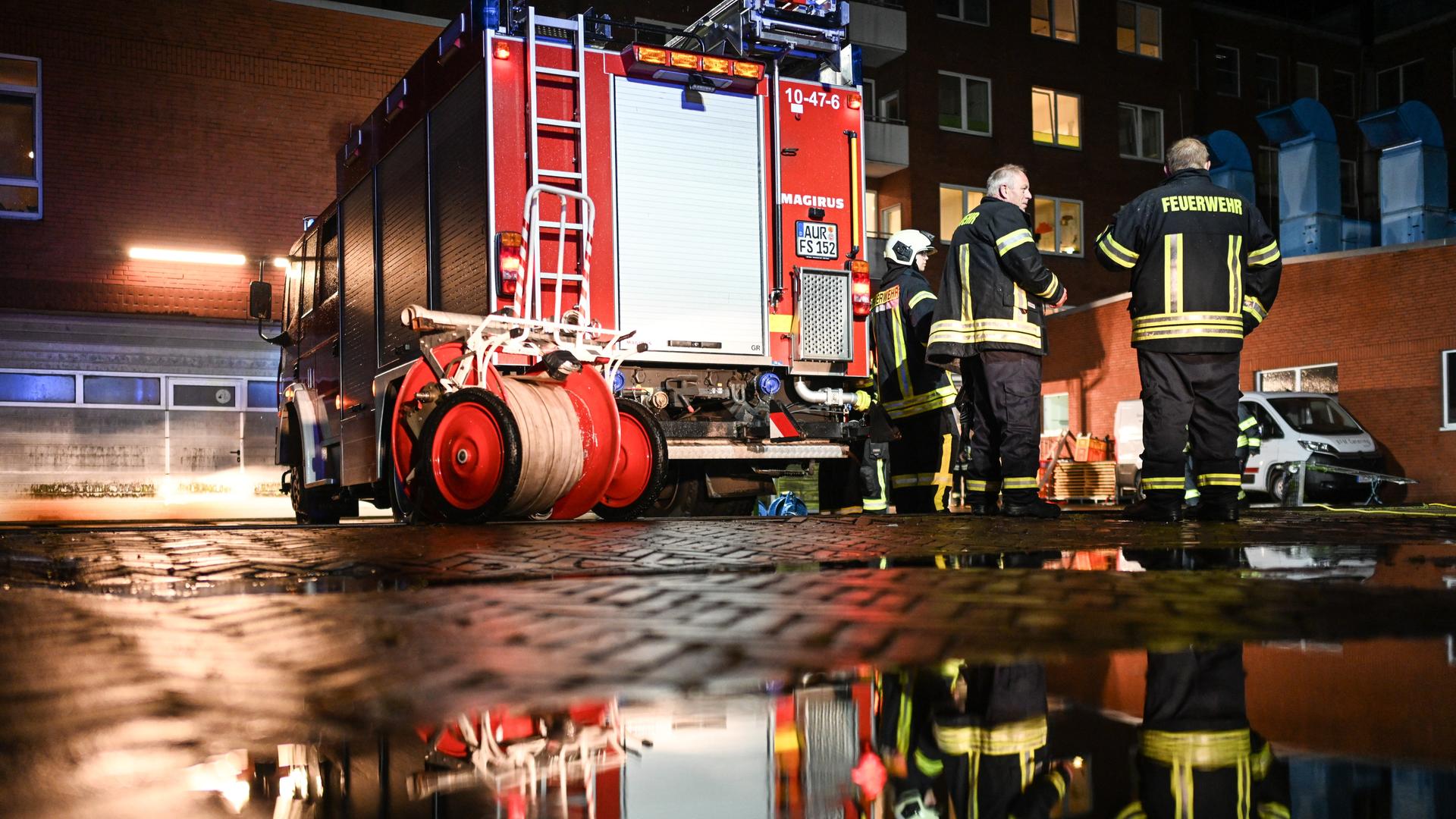 Es ist dunkel. Ein Feuerwehrauto spiegelt sich in einer Pfütze. Daneben stehen Feuerwehrleute. Im Hintergrund das Krankenhaus mit beleuchteten Fenstern.