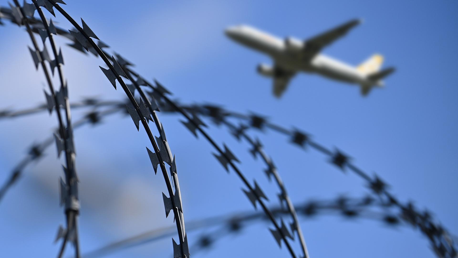Ein startendes Passagierflugzeug ist am Himmel zu sehen, im Vordergrund ein Zaun mit Stacheldraht. Ein startendes Passagierflugzeug ist am Himmel zu sehen, im Vordergrund ein Zaun mit Stacheldraht.