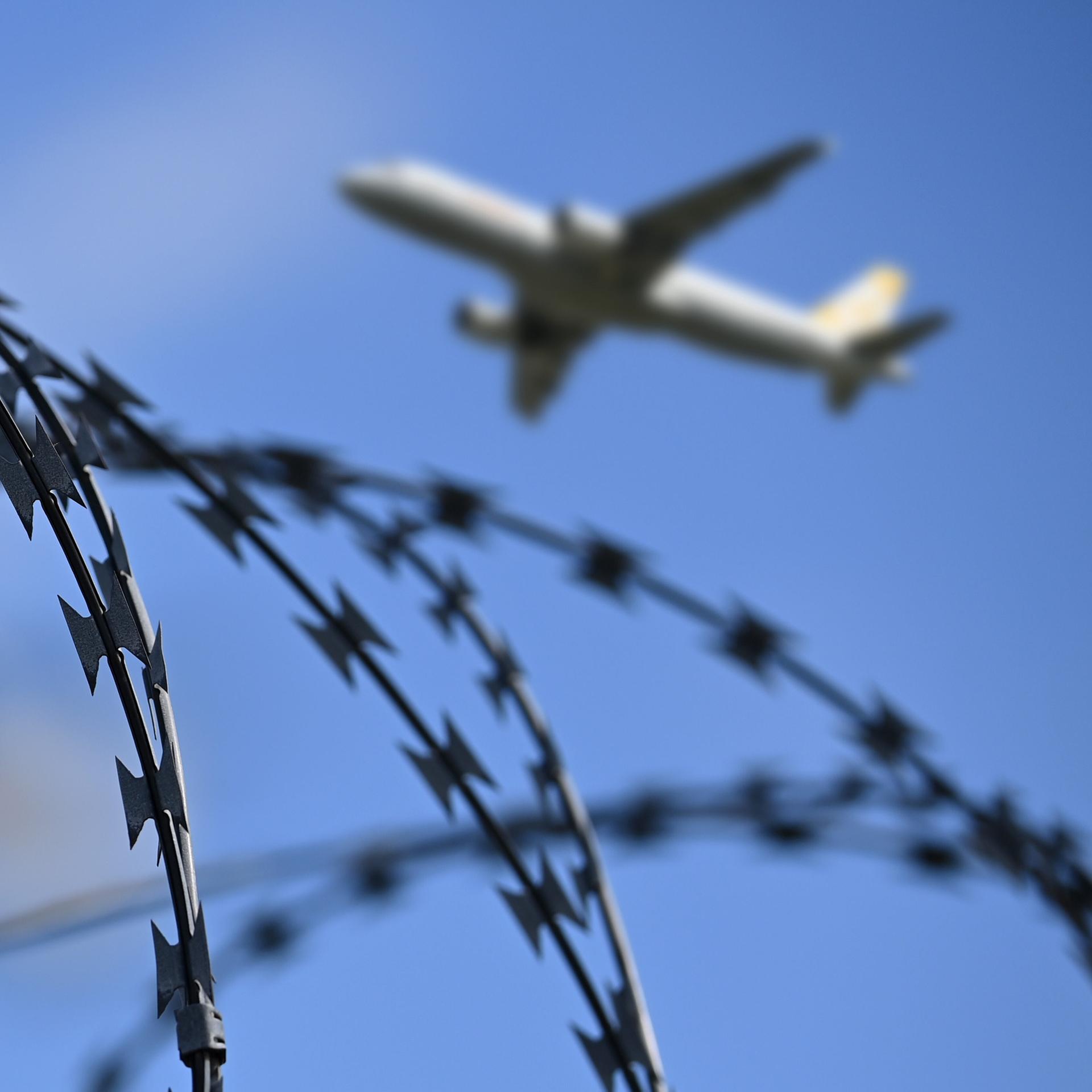Ein startendes Passagierflugzeug ist am Himmel zu sehen, im Vordergrund ein Zaun mit Stacheldraht.
