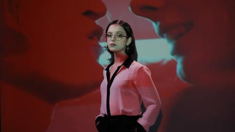 Elizabeth Duval, mit Brille und Kreolen, in einer pinken Bluse und schwarzer Hose, steht vor der Projektion eines Filmausschnitts, in dem zwei Münder einander fast berühren.