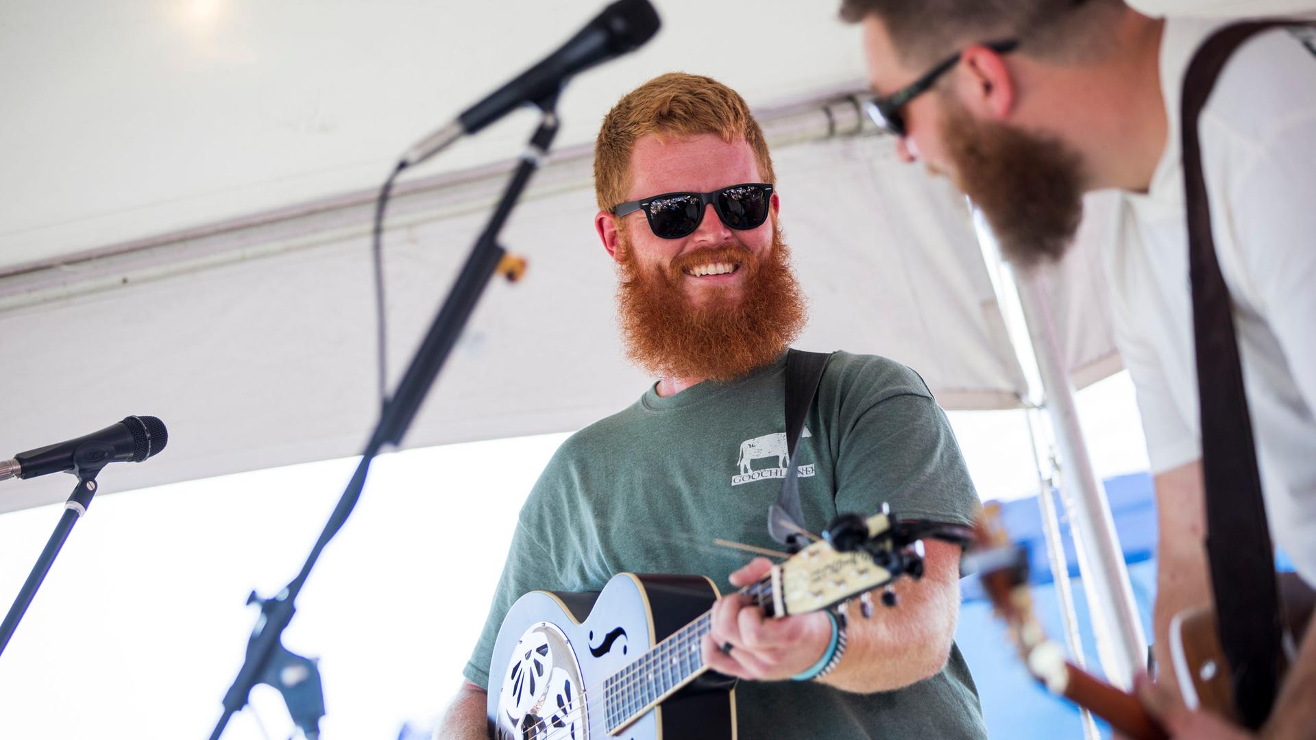Ein Mann mit einem roten Vollbart trägt ein grünes T-Shirt und eine schwarze Sonnenbrille, er spielt Gitarre und schaut lächelnd zu einem Mitmusiker: Es handelt sich um Oliver Anthony bei einem Auftritt in Moyock in North Carolina am 19. August 2023.