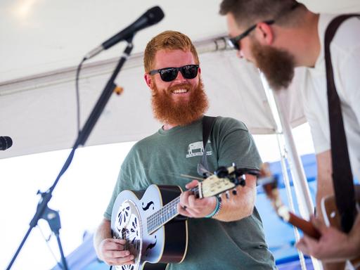 Ein Mann mit einem roten Vollbart trägt ein grünes T-Shirt und eine schwarze Sonnenbrille, er spielt Gitarre und schaut lächelnd zu einem Mitmusiker: Es handelt sich um Oliver Anthony bei einem Auftritt in Moyock in North Carolina am 19. August 2023.