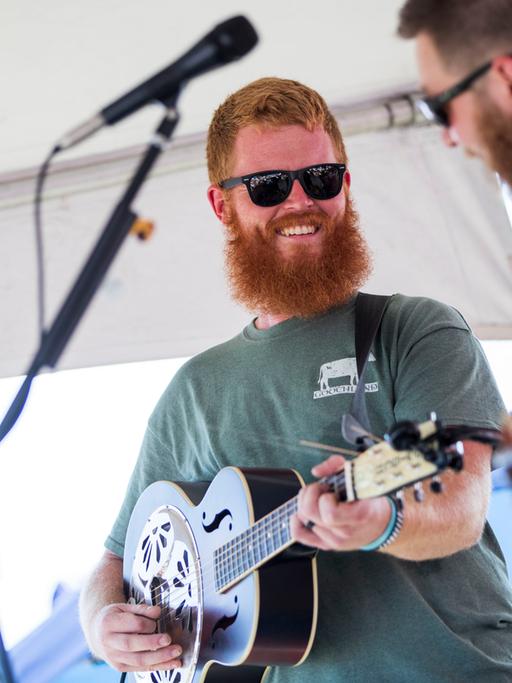 Ein Mann mit einem roten Vollbart trägt ein grünes T-Shirt und eine schwarze Sonnenbrille, er spielt Gitarre und schaut lächelnd zu einem Mitmusiker: Es handelt sich um Oliver Anthony bei einem Auftritt in Moyock in North Carolina am 19. August 2023.