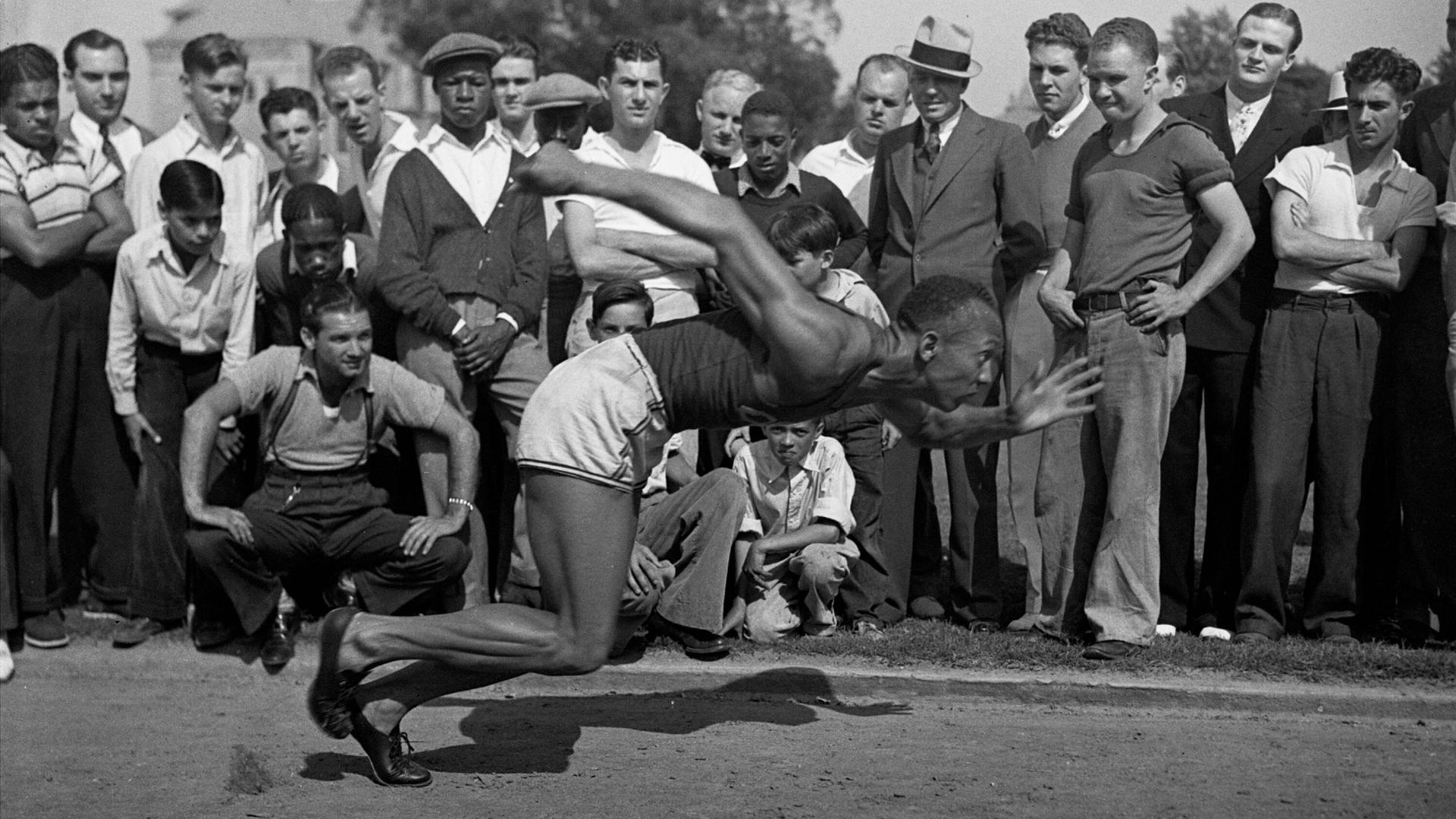 Ein Schwarz-Weiß-Foto zeigt Jesse Owens in einer dynamischen Pose beim Training. Eine Gruppe von Zuschauern beobachtet ihn dabei. Ein Schwarz-Weiß-Foto zeigt Jesse Owens in einer dynamischen Pose beim Training. Eine Gruppe von Zuschauern beobachtet ihn dabei.