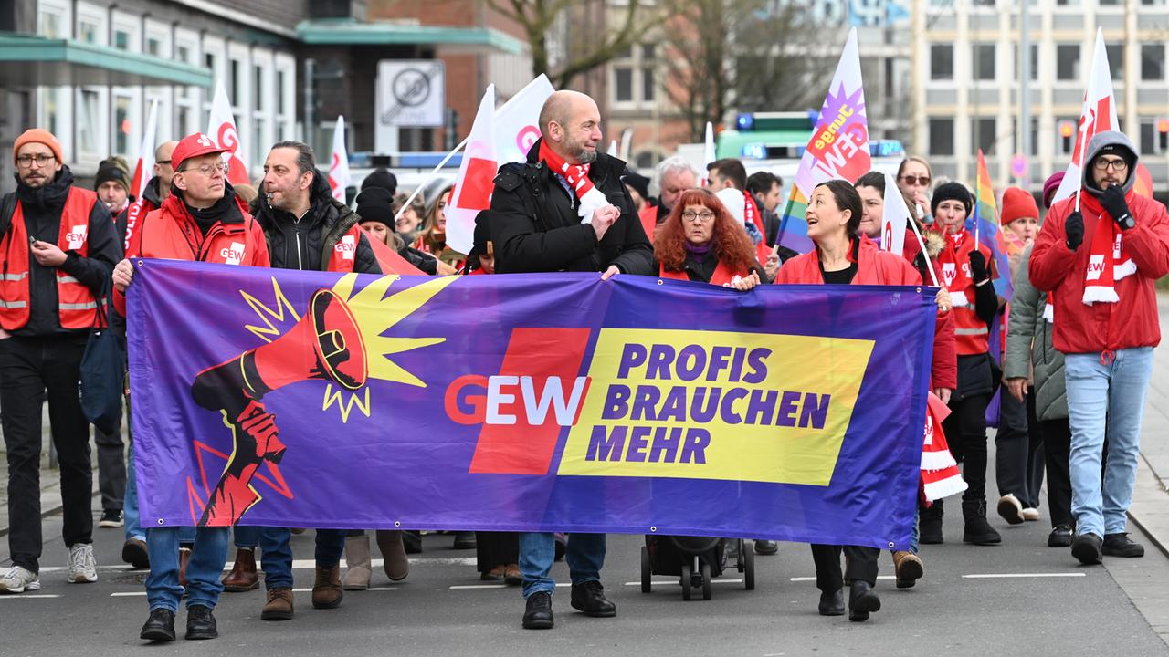 Hinter einem Banner der Gewerkschaft Erziehung und Wissenschaft laufen mehrere Personen, die an einem Streik teilnehmen. Auf dem Banner steht "Profis brauchen mehr".