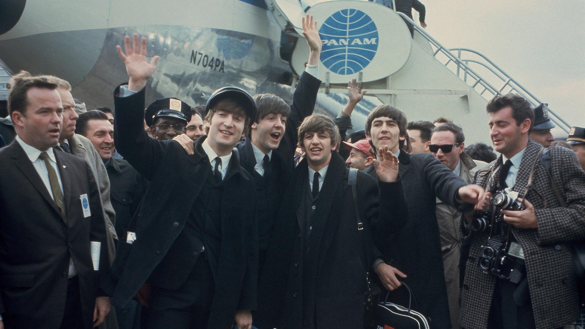 John Lennon winkend, Paul McCartney, Ringo Starr and George Harrison stehen vor einem Jumbo (AP Photo), im Hintergrund die Treppe, die sie vom Flugzeug herabgegangen sind. Die vier Musiker sind umgeben von Männern, rechts steht ein Fotograf.
