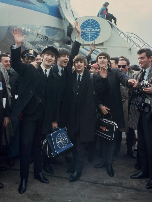 John Lennon winkend, Paul McCartney, Ringo Starr and George Harrison stehen vor einem Jumbo (AP Photo), im Hintergrund die Treppe, die sie vom Flugzeug herabgegangen sind. Die vier Musiker sind umgeben von Männern, rechts steht ein Fotograf.