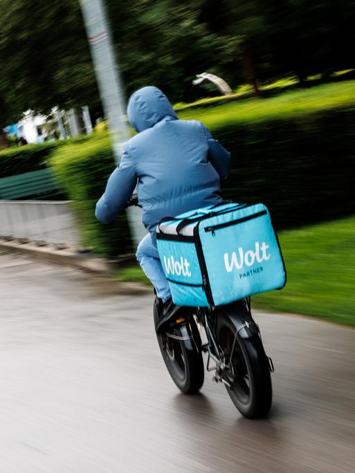 Ein Fahrradkurier von Wolt fährt im Anorak bei Herbstwetter eine Straße entlang. Ein Fahrradkurier von Wolt fährt im Anorak bei Herbstwetter eine Straße entlang.
