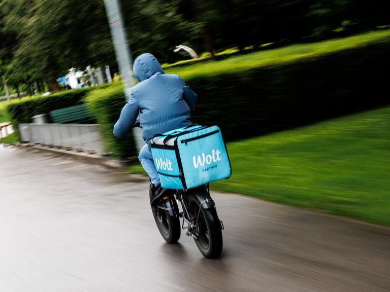 Ein Fahrradkurier von Wolt fährt im Anorak bei Herbstwetter eine Straße entlang. Ein Fahrradkurier von Wolt fährt im Anorak bei Herbstwetter eine Straße entlang.