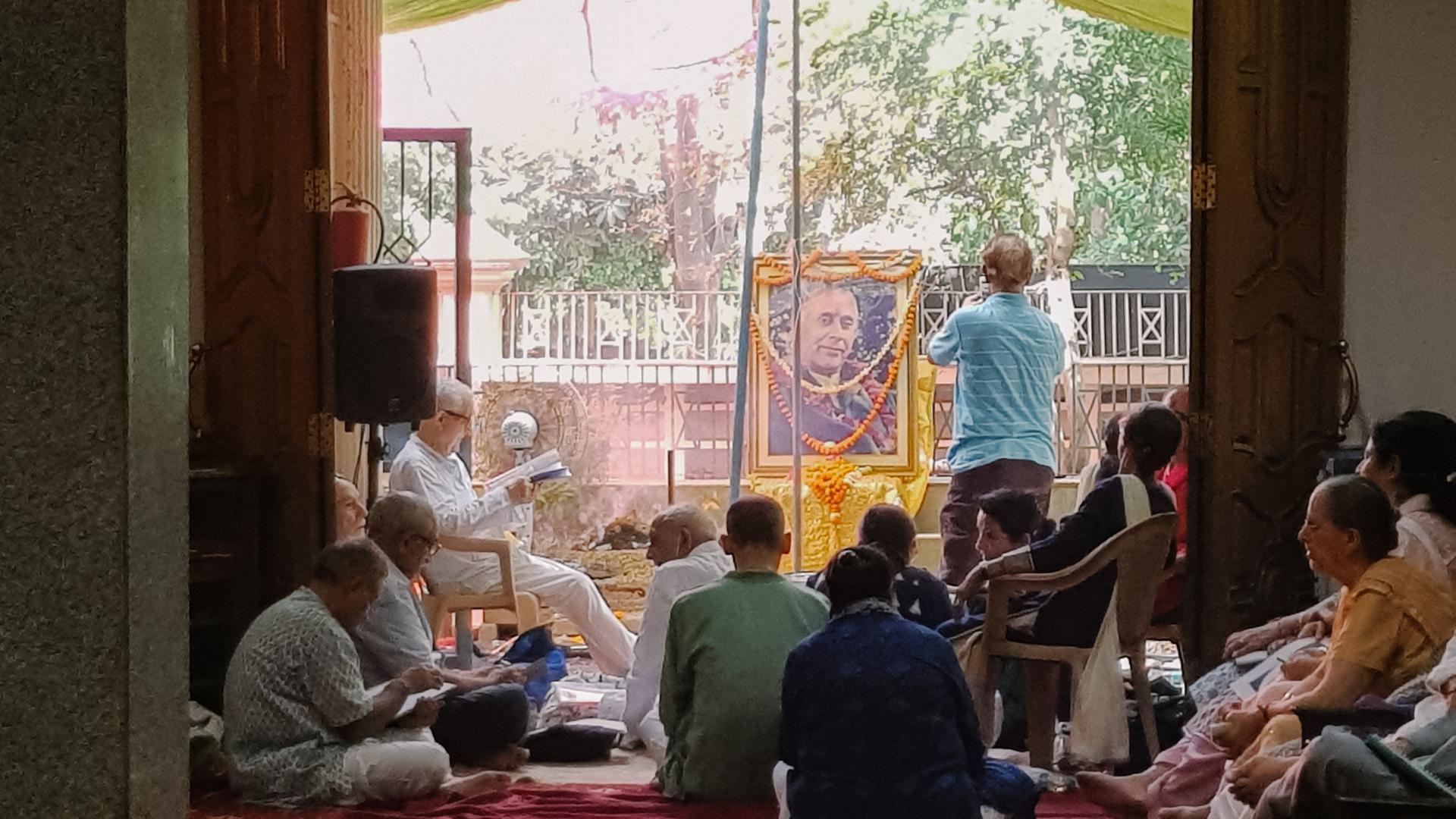 Menschen sitzen und knien in einem hindustischen Tempel vor dem golden gerahmten und mit Blumengirlanden geschmückten Porträt eines Mannes.