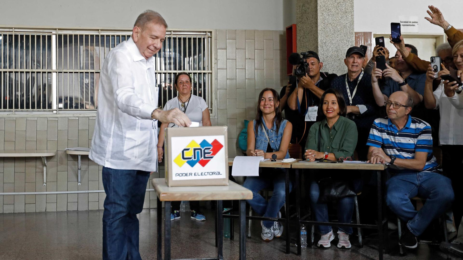 González Urrutia steht an einer Urne in einem Gebäude und wirft seinen Stimmzettel ein. An einem Tisch sitzen Wahlhelfer. Am rechten Bildrand viele Journalisten mit Handys und Kameras.