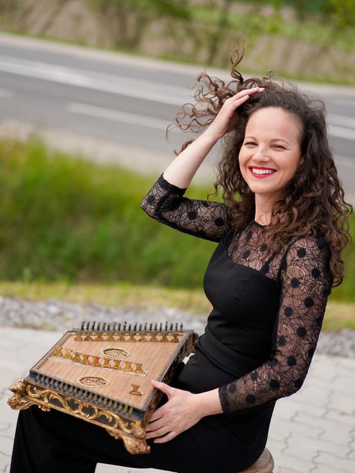 Franziska Fleischanderl, eine Frau mit lockigem, braunem Haar, sitzt am Rande einer Straße. Sie hat ein historisches Hackbrett auf dem Schoß. Sie ist schwarz gekleidet und lacht in die Kamera.