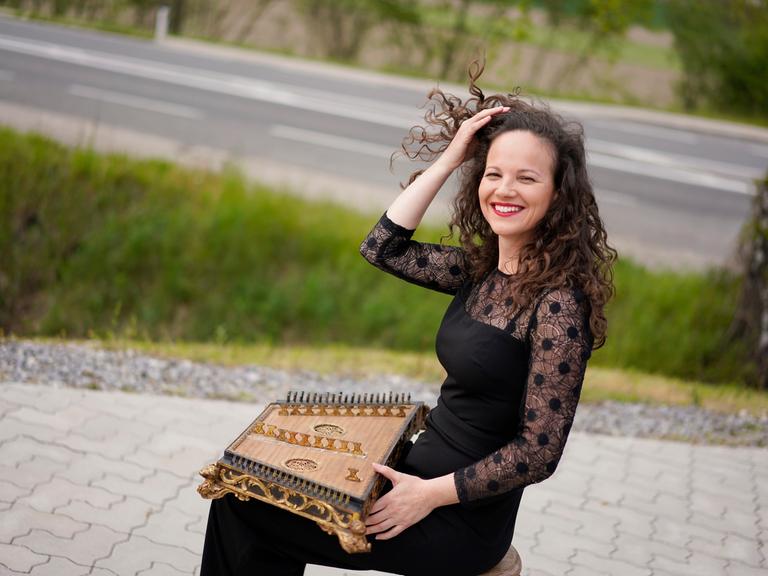 Franziska Fleischanderl, eine Frau mit lockigem, braunem Haar, sitzt am Rande einer Straße. Sie hat ein historisches Hackbrett auf dem Schoß. Sie ist schwarz gekleidet und lacht in die Kamera.