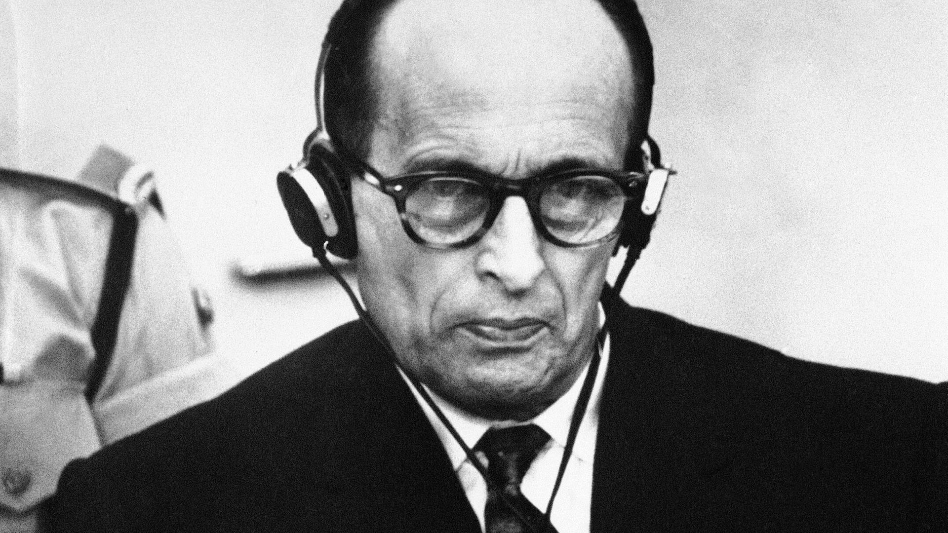 Adolf Eichmann sitzt mit ernster Miene vor Gericht. Er trägt einen Kopfhörer.