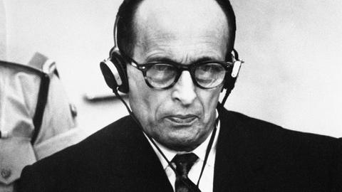 Adolf Eichmann sitzt mit ernster Miene vor Gericht. Er trägt einen Kopfhörer.