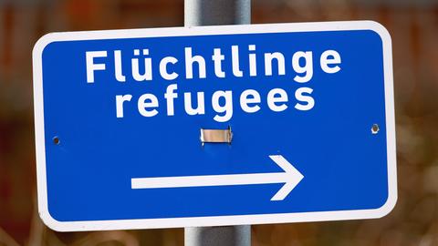 Ein kleines blaues, schief hängendes Hinweisschild mit der Aufschrift "Flüchtlinge refugees" und einem Pfeil nach rechts an einem Schildermast.