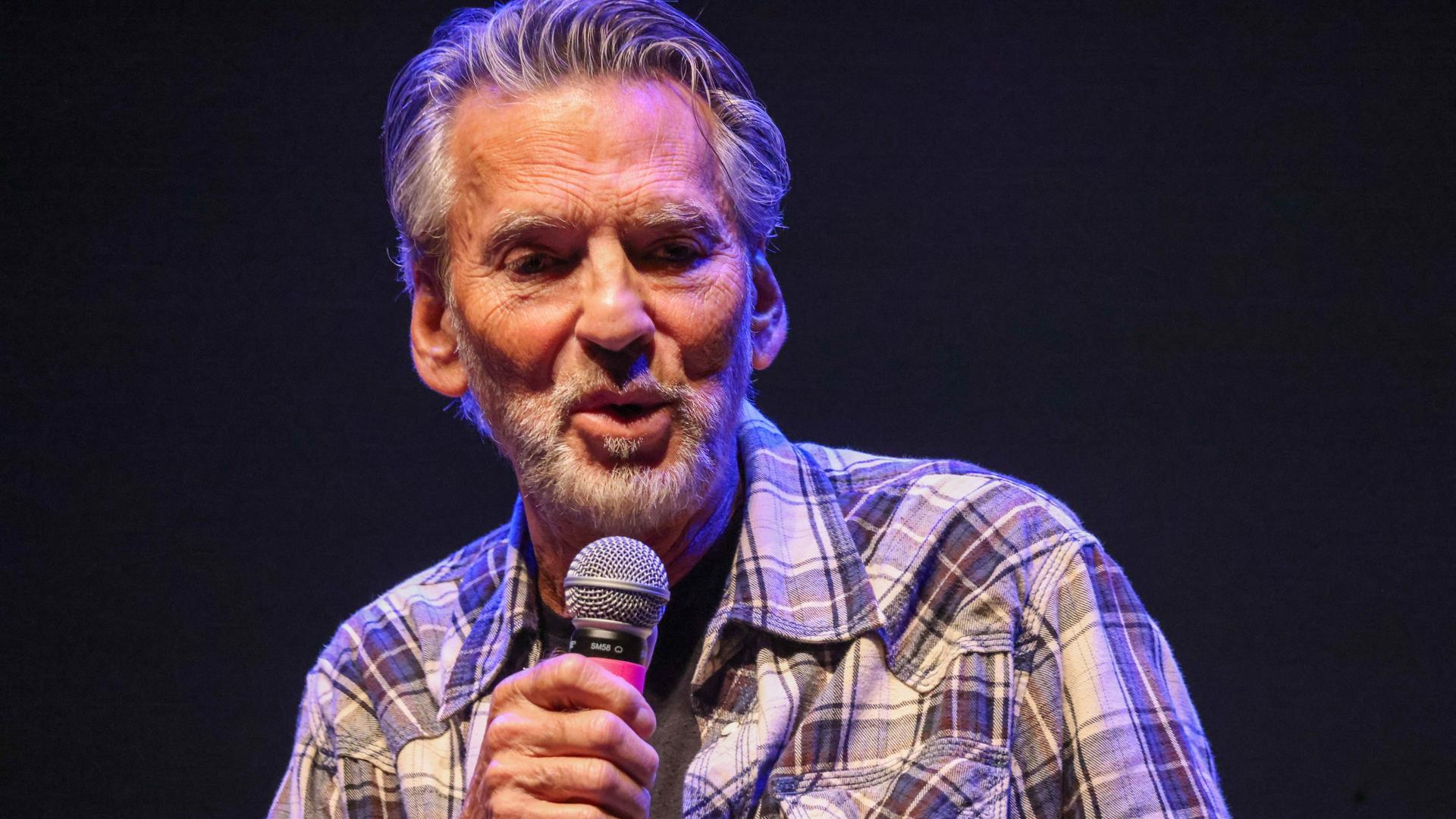 Kenny Loggins singt mit Mikro in der Hand auf einer Bühne. Der Hintergrund ist dunkel. Kenny Loggins singt mit Mikro in der Hand auf einer Bühne. Der Hintergrund ist dunkel.